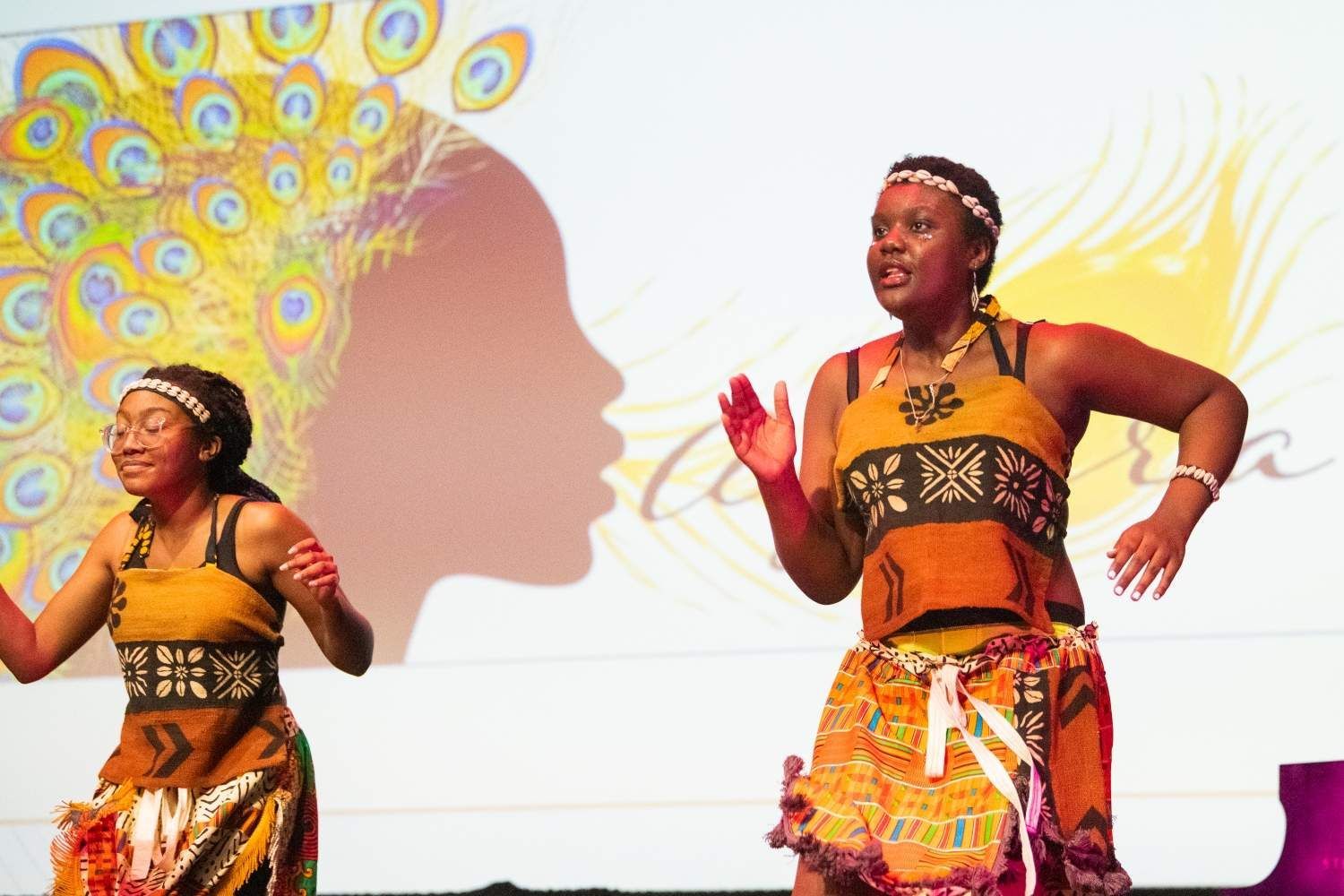 Two women in traditional African clothing dance on stage, colorful backdrop.