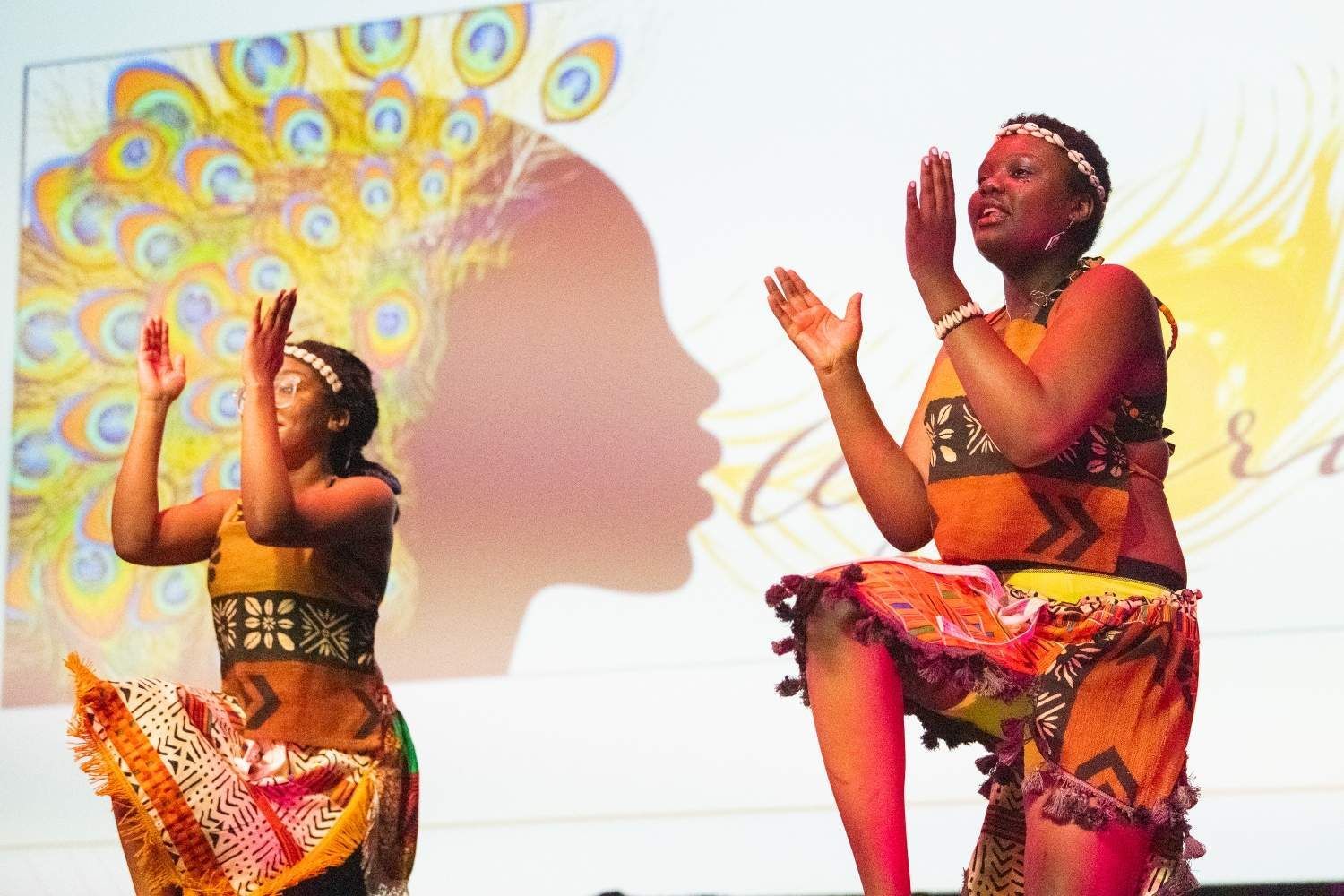 Two women in traditional African outfits dance on stage, hands raised, with a backdrop of a peacock and a woman's silhouette.