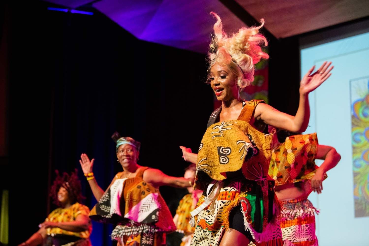 African dancers in colorful traditional clothing perform on stage, smiling and raising their arms.