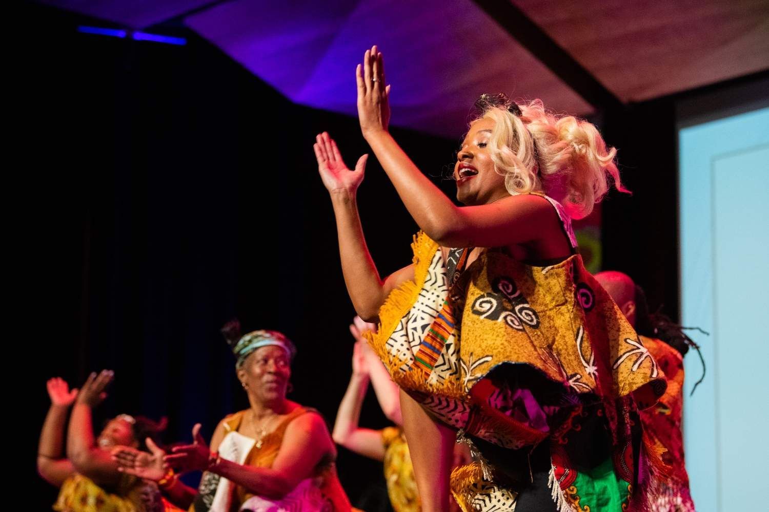 Woman in colorful African print dress dances onstage with arms raised. Other women clap in the background.