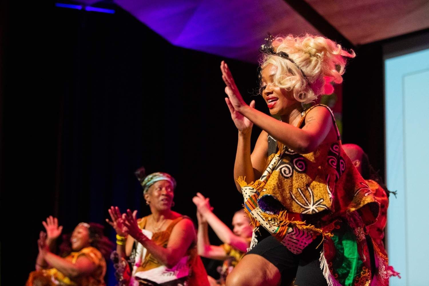 Dancers in vibrant African-print outfits clap onstage, lit by blue and purple lights. Blonde dancer in focus.
