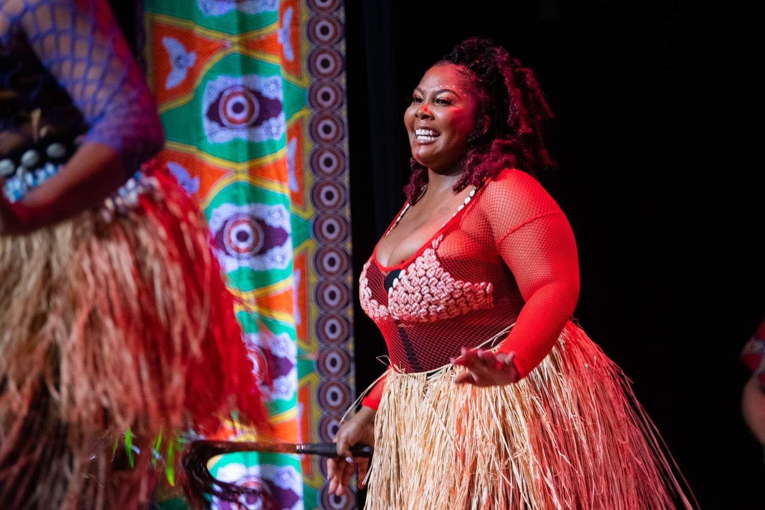 Woman in red mesh and grass skirt smiles while dancing onstage.