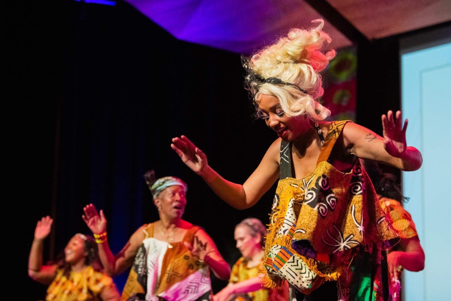 Dancers in colorful African print dresses perform onstage, arms raised. Woman with blonde updo smiles.