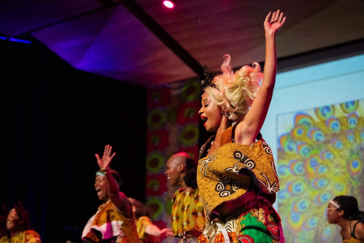 Dancers on stage in colorful African-inspired costumes, arms raised. A projector screen shows a peacock design.