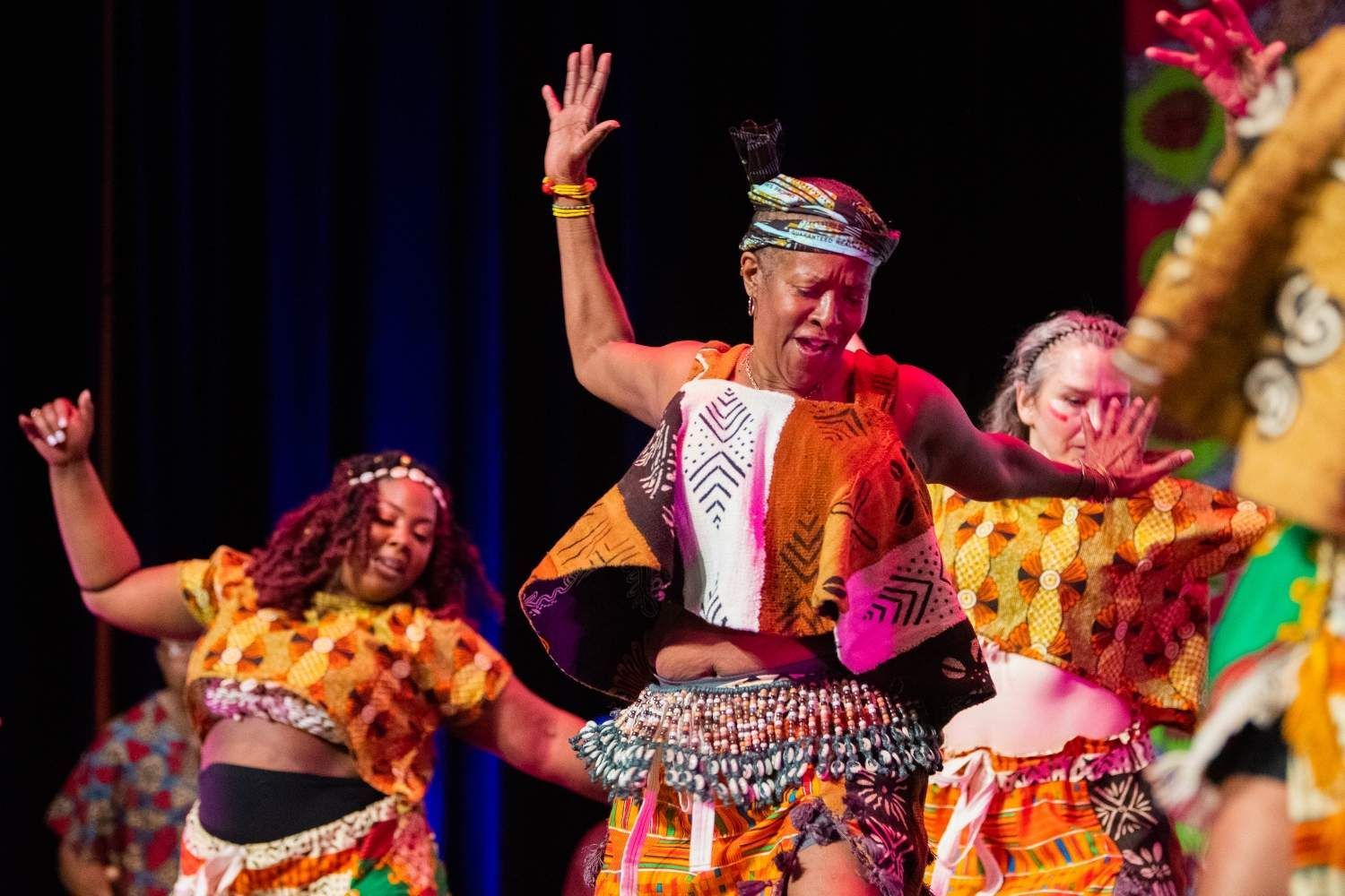 Dancers in colorful African-style attire perform onstage, raising arms in a joyful expression.