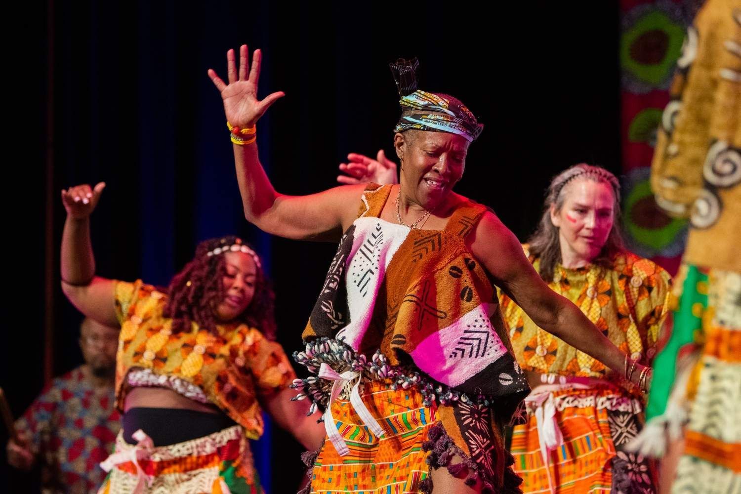 People in colorful African attire dance on a stage, raising their arms joyfully.