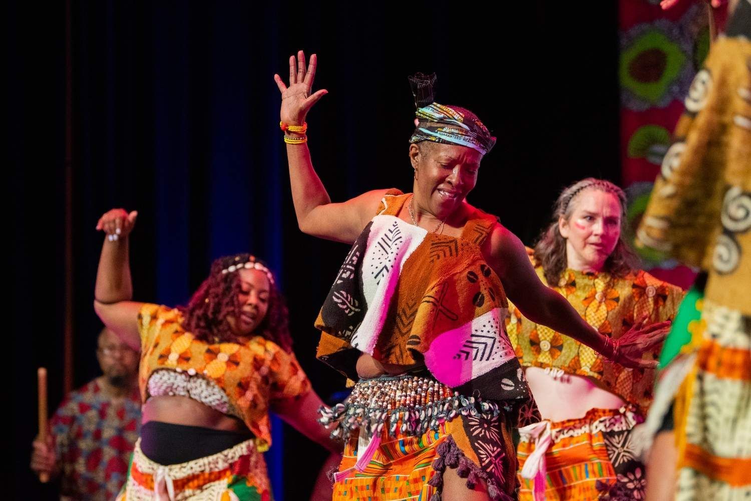 Dancers in colorful African attire perform on stage, joyful expressions, arms raised.