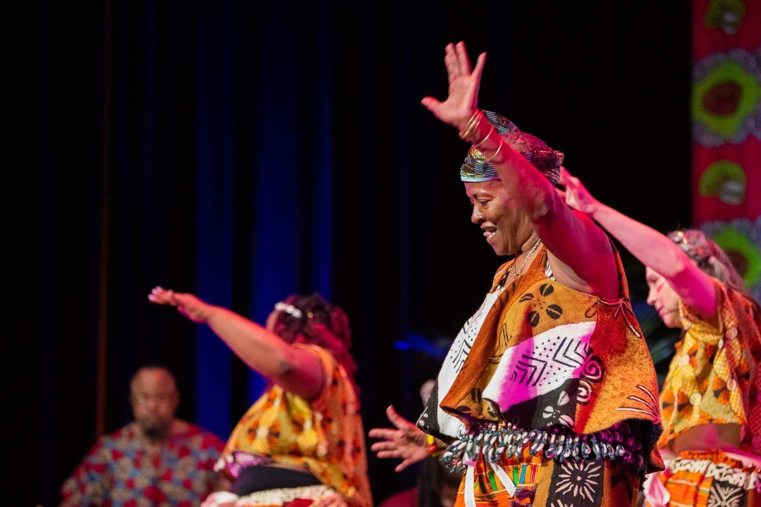 African dancers in vibrant outfits perform on stage, raising arms with joyous expressions.