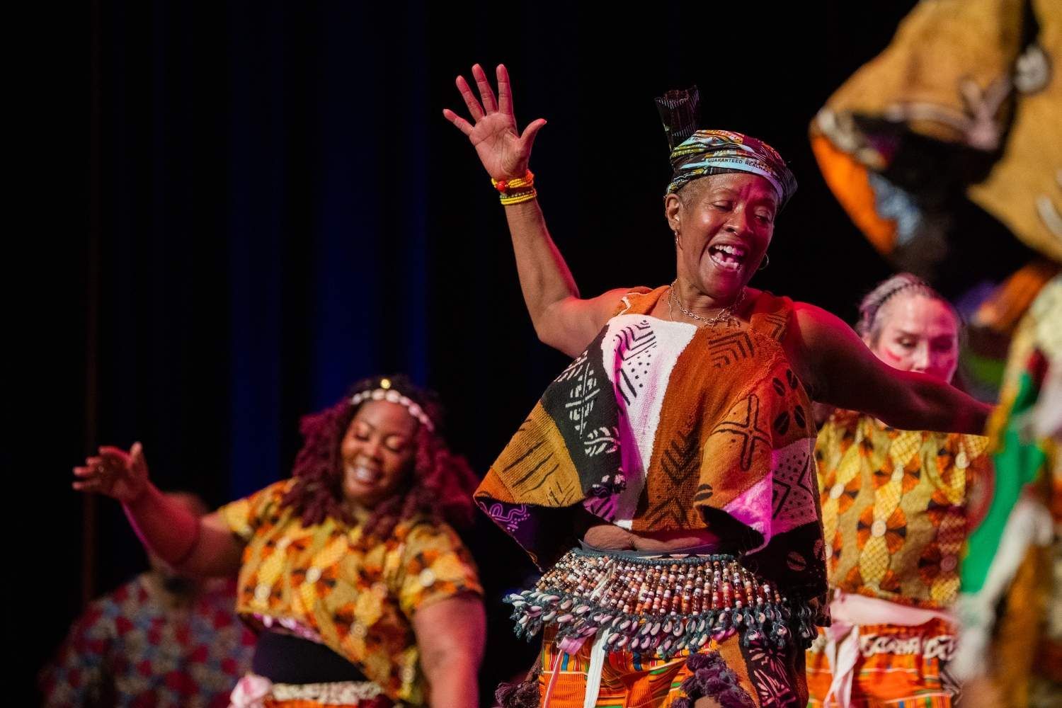 People in colorful African attire dance on stage, raising arms and smiling.