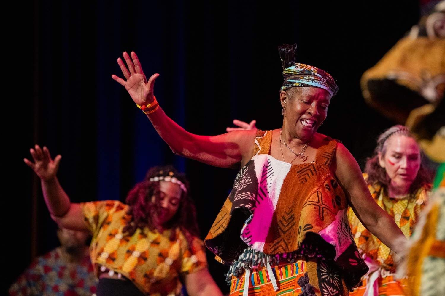Dancers in colorful African attire performing on stage. Woman in focus with arms raised, smiling.