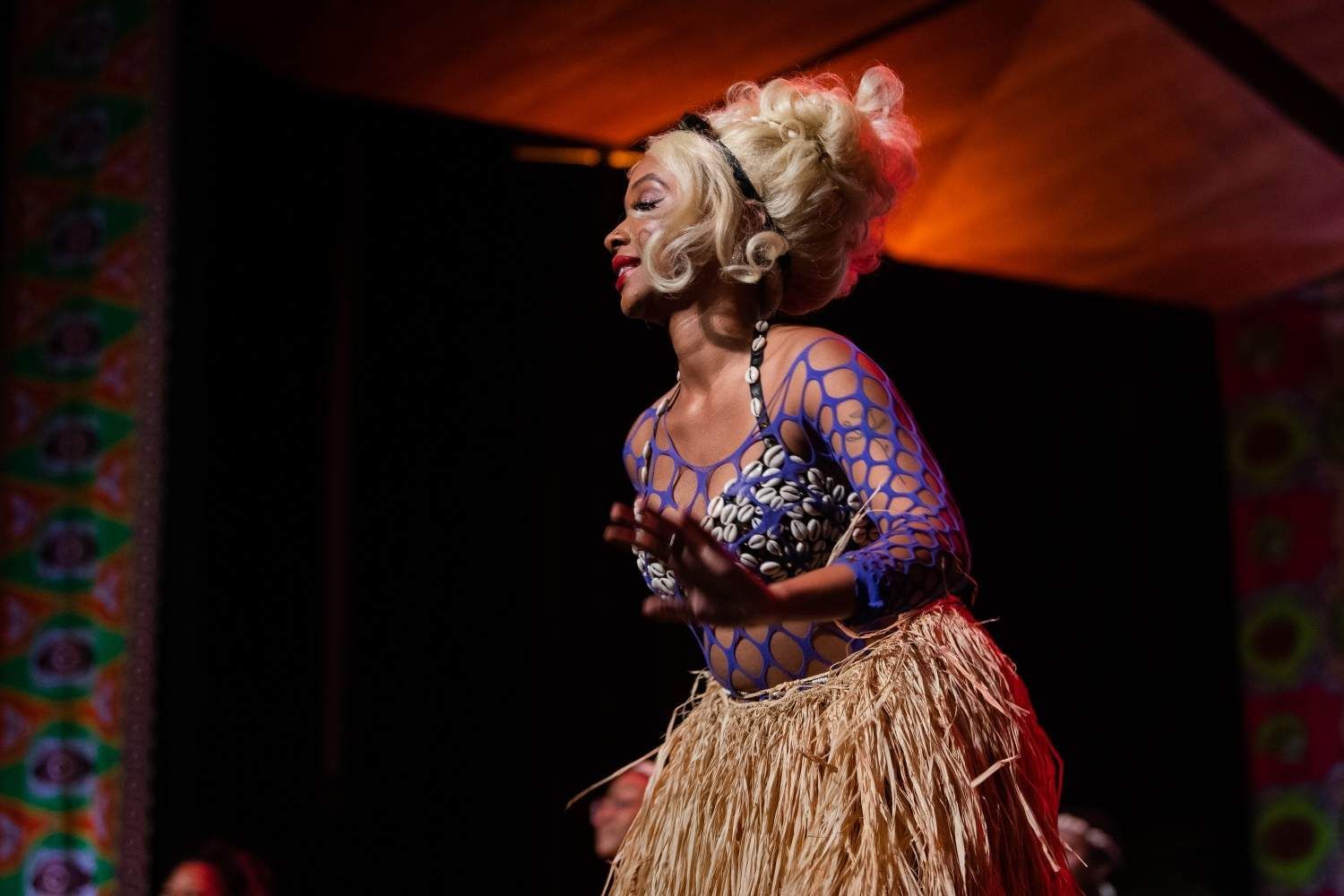 Woman in a costume, performing on stage. Blonde hair, blue top, straw skirt, colorful backdrop.