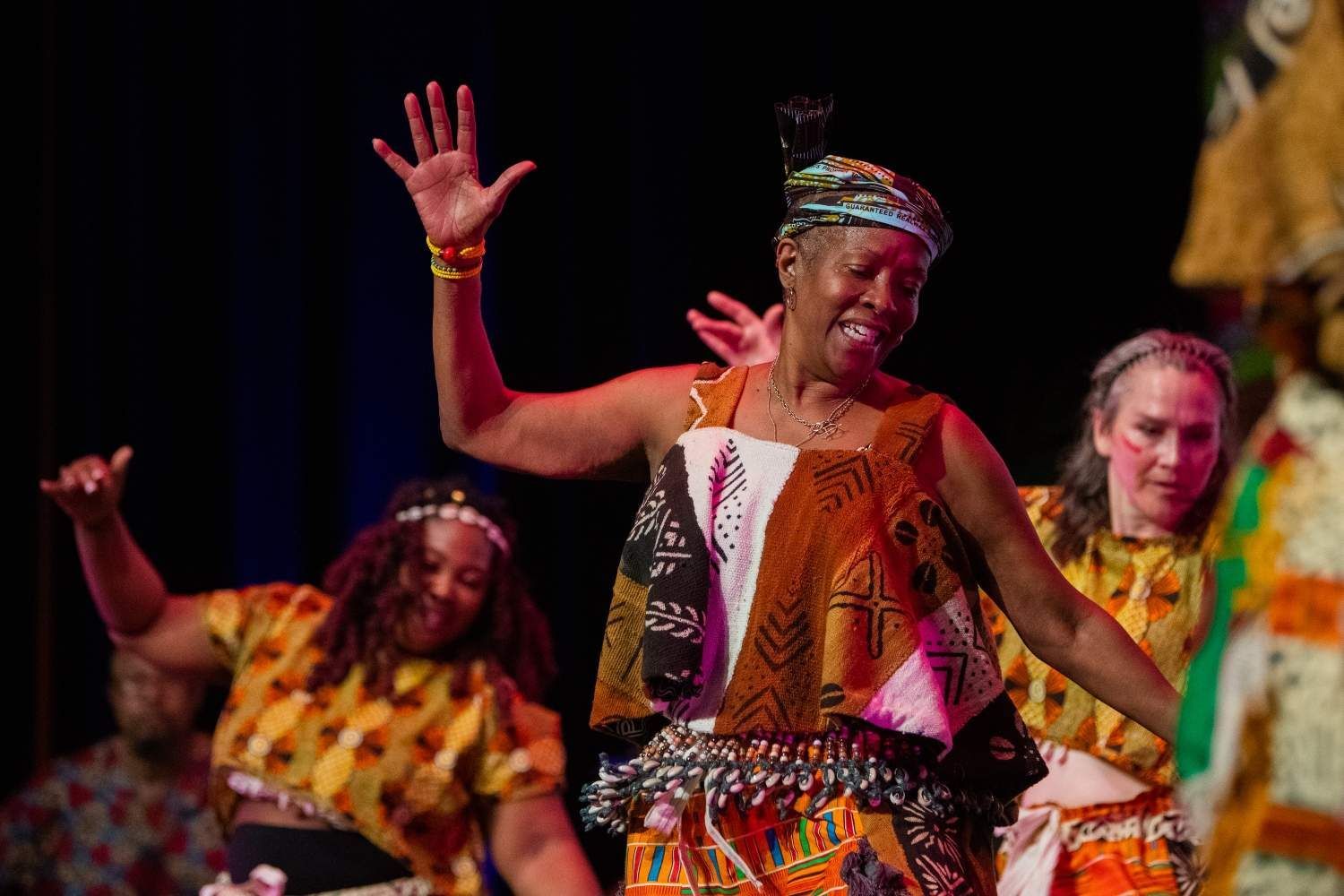 Three women dancing on stage, wearing colorful African-inspired clothing. They raise their arms and smile.