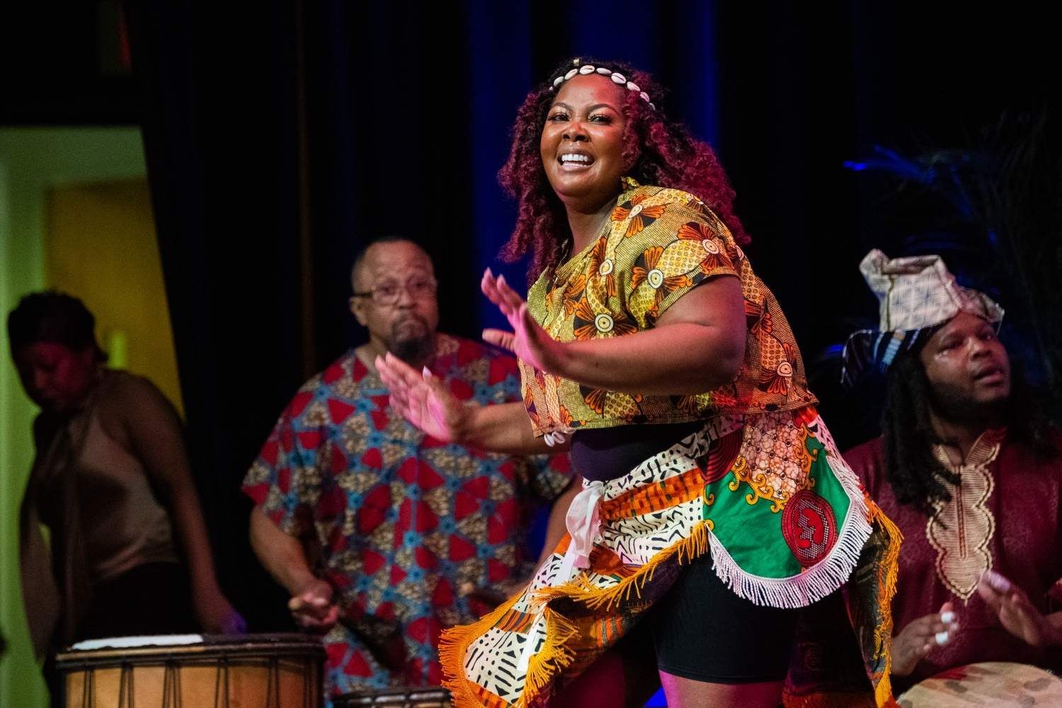 Woman dancing onstage in African-inspired attire; musicians in background, playing drums.