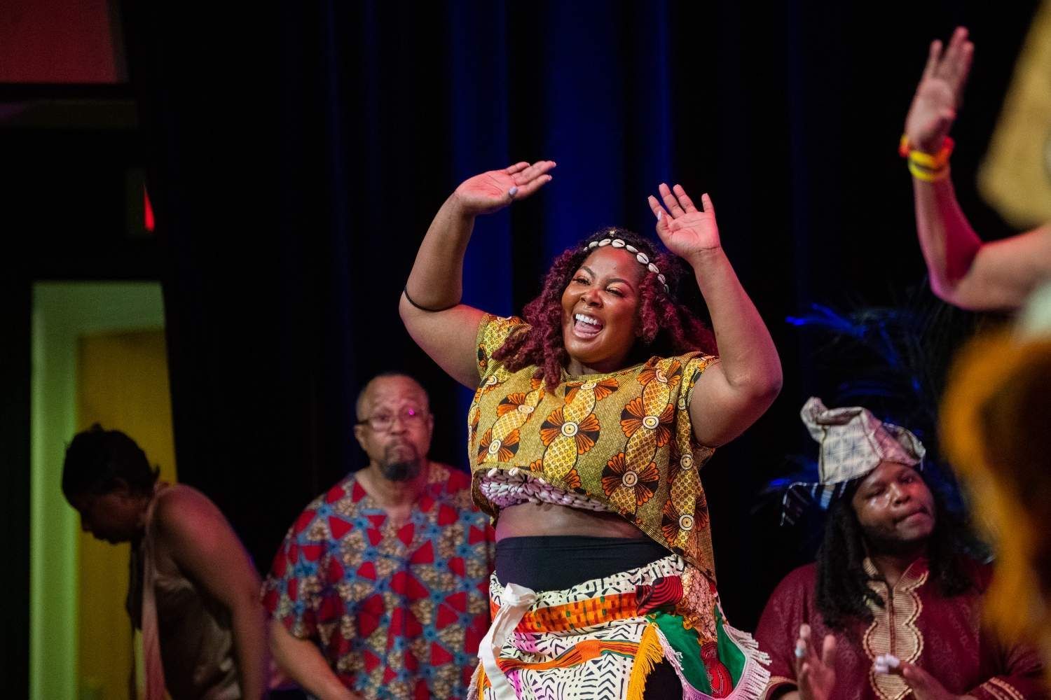 Woman in African print top dancing, arms raised, on stage with other performers.