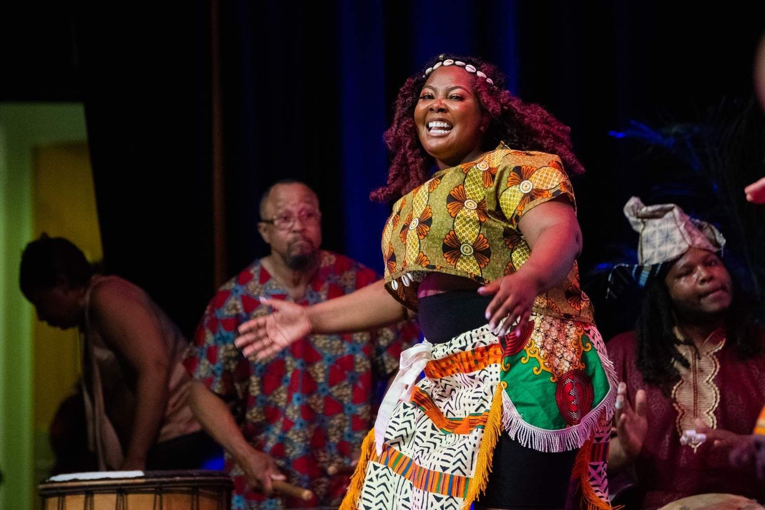 Woman in colorful African attire sings and dances on stage with musicians.
