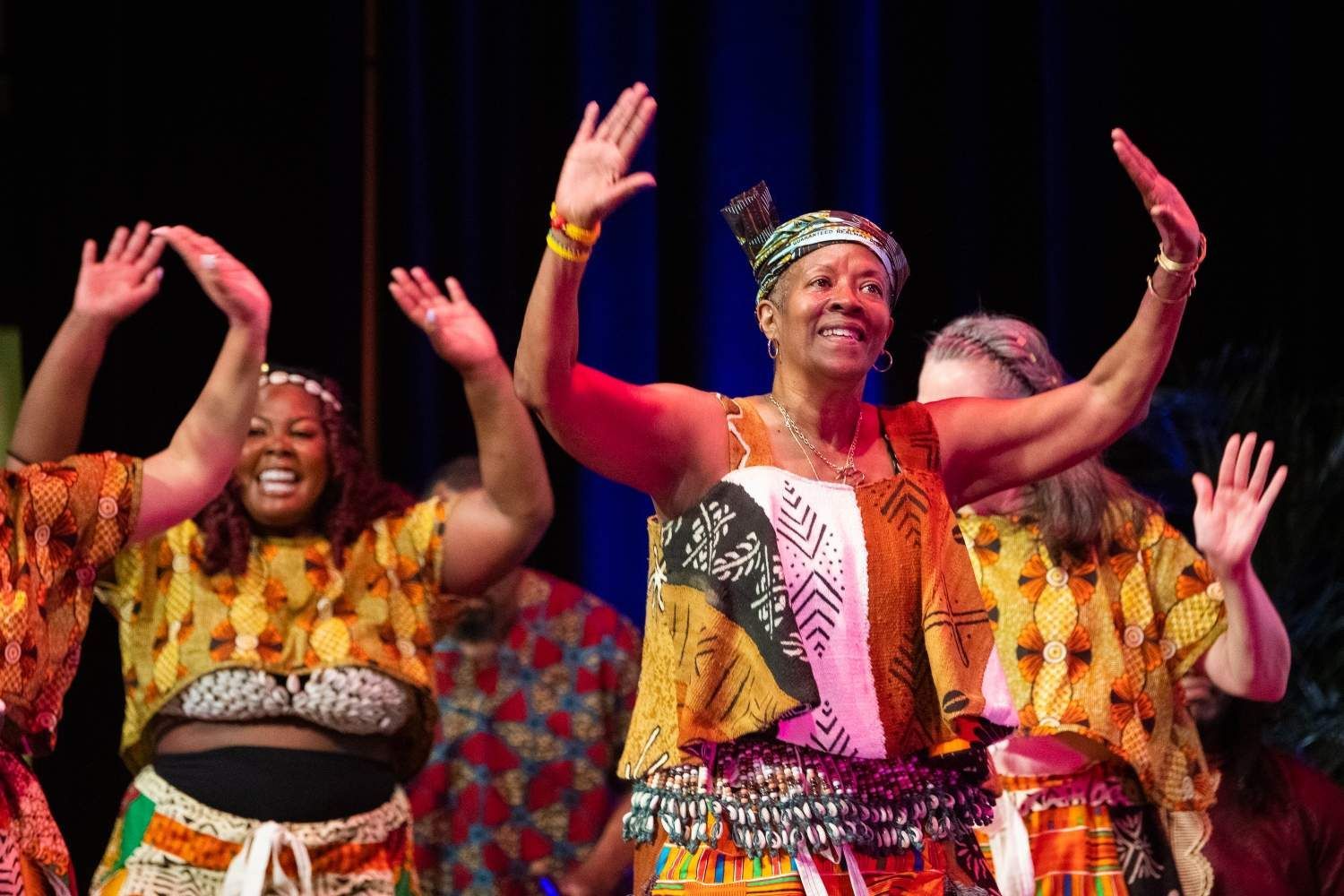 Group of women dancing on stage, arms raised, wearing colorful African-print clothing.