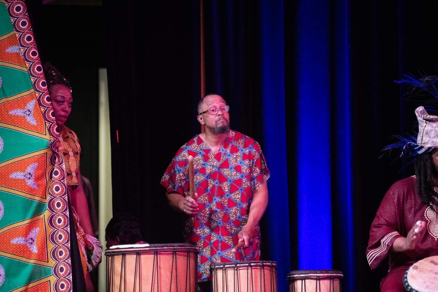 Three people drumming on stage, wearing colorful African-inspired clothing.