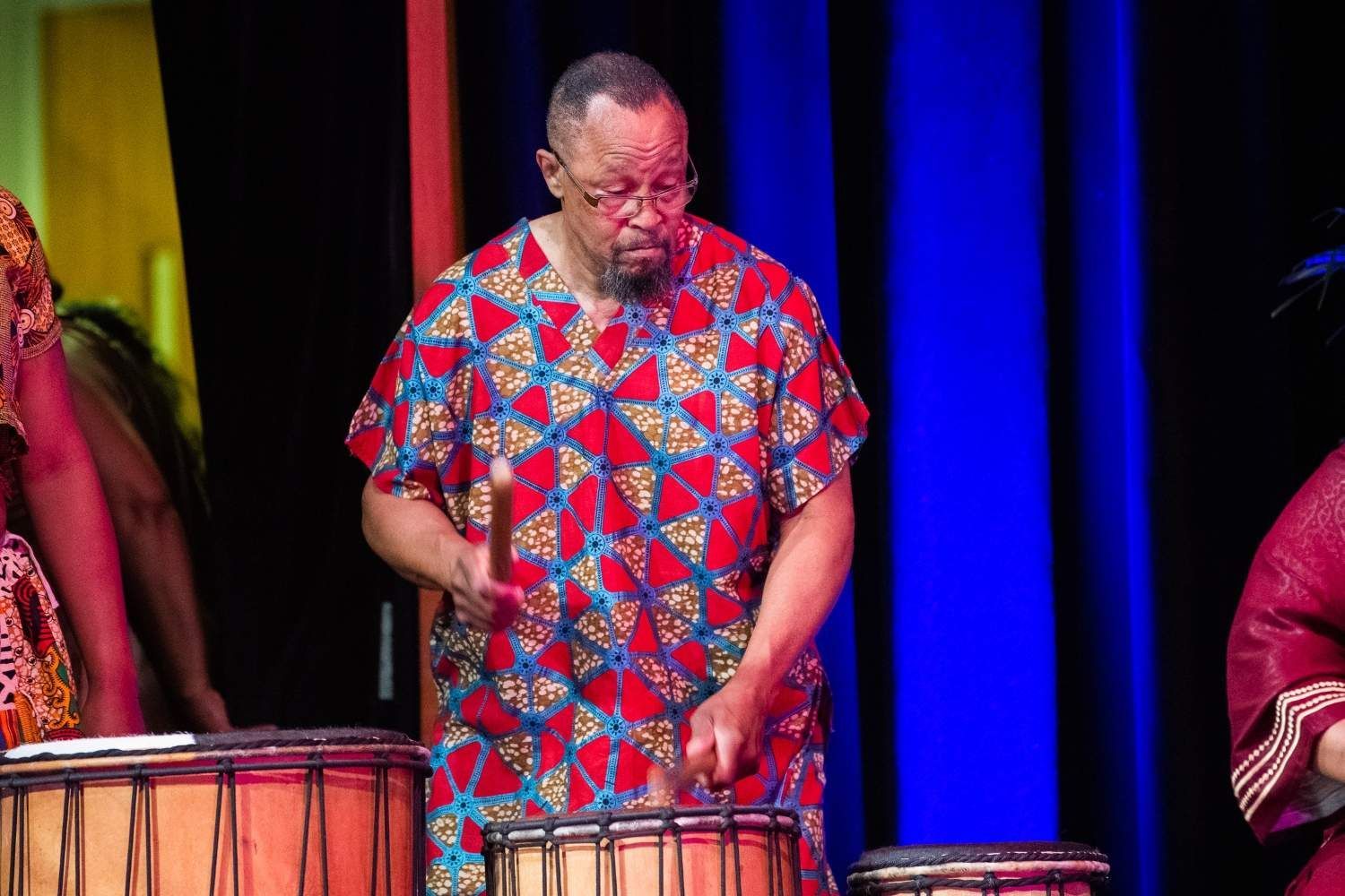 Man in patterned red and blue shirt playing drums on stage.