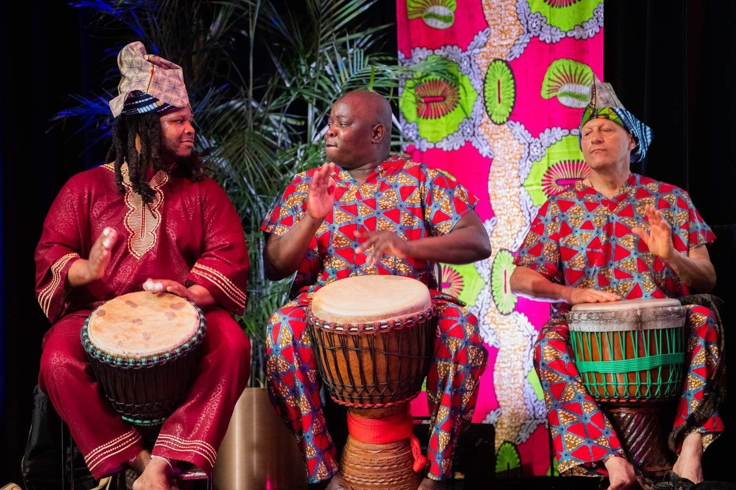 Three people in vibrant African attire playing drums, smiling, indoors.
