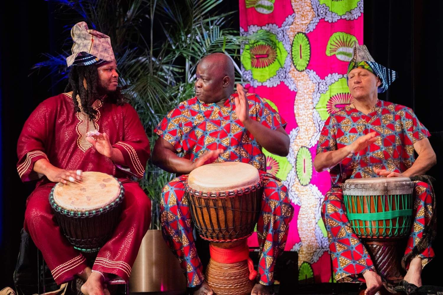 Three people playing drums, dressed in bright patterned outfits. Dark setting, palm fronds.