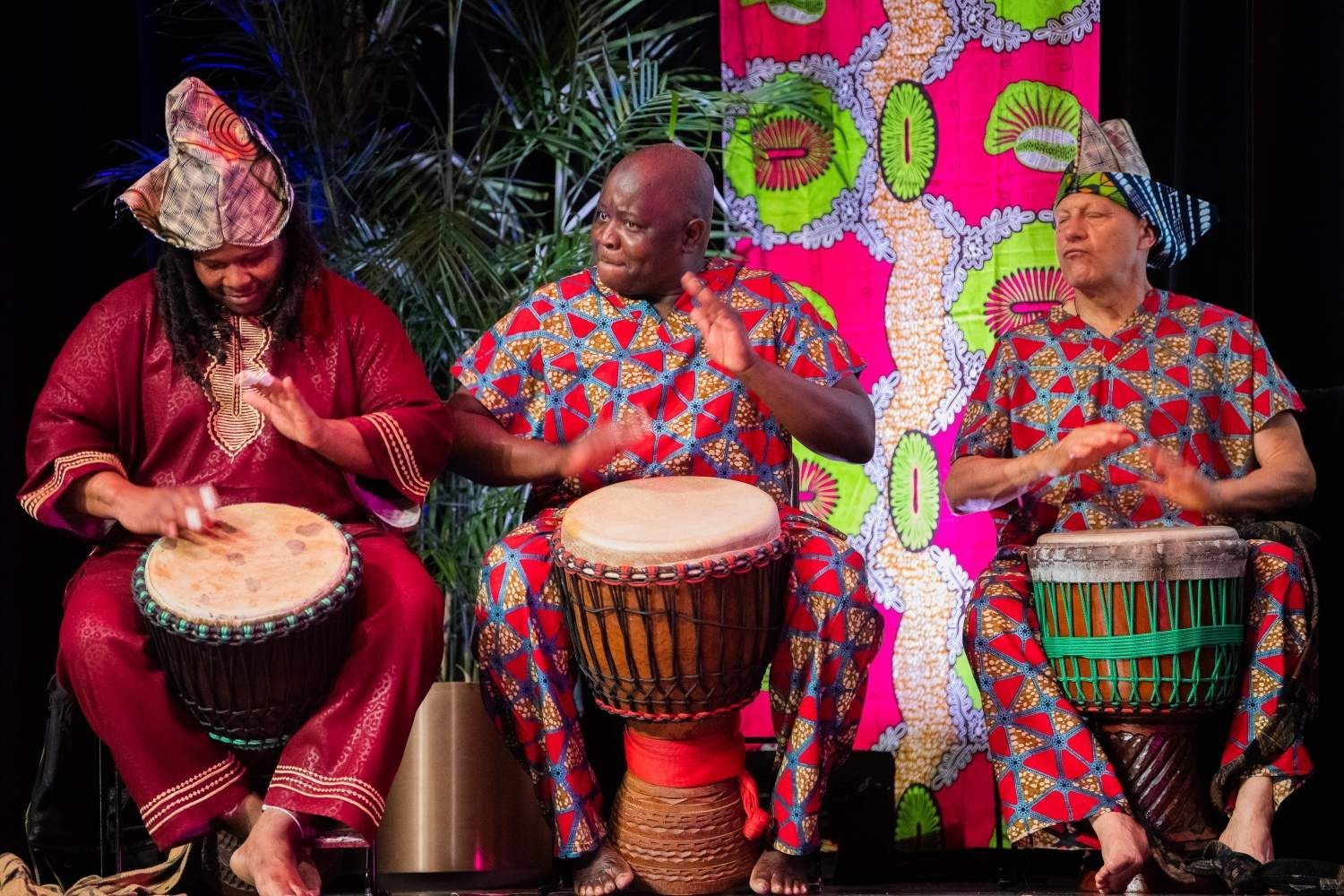 Three people in African-print clothing playing drums. Stage setting with bright backdrop.
