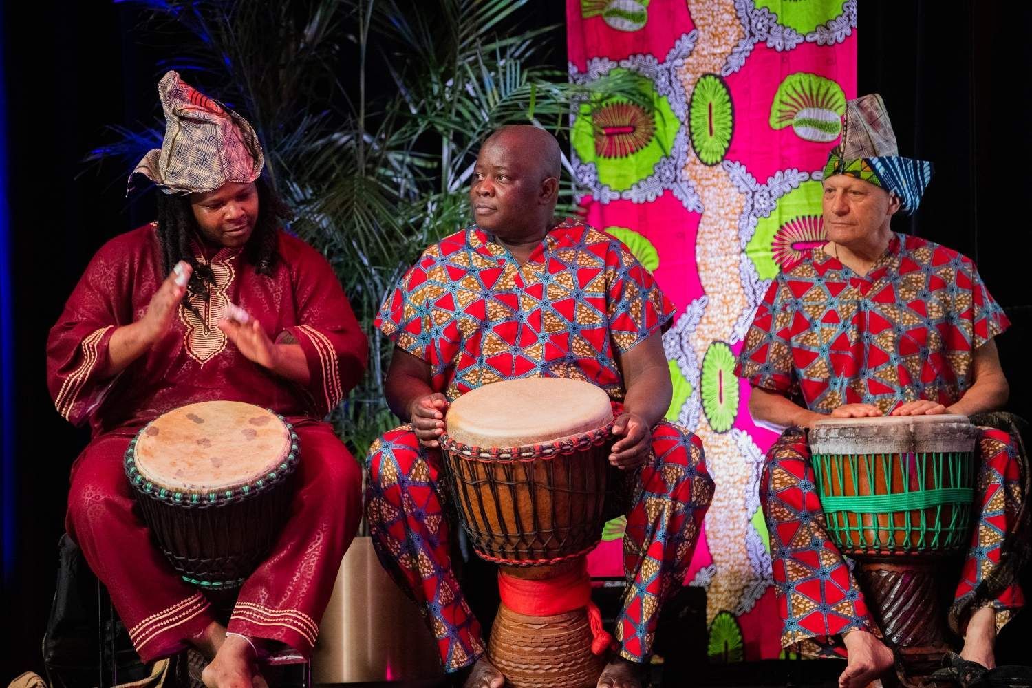 Three people in patterned African attire playing djembe drums on stage.