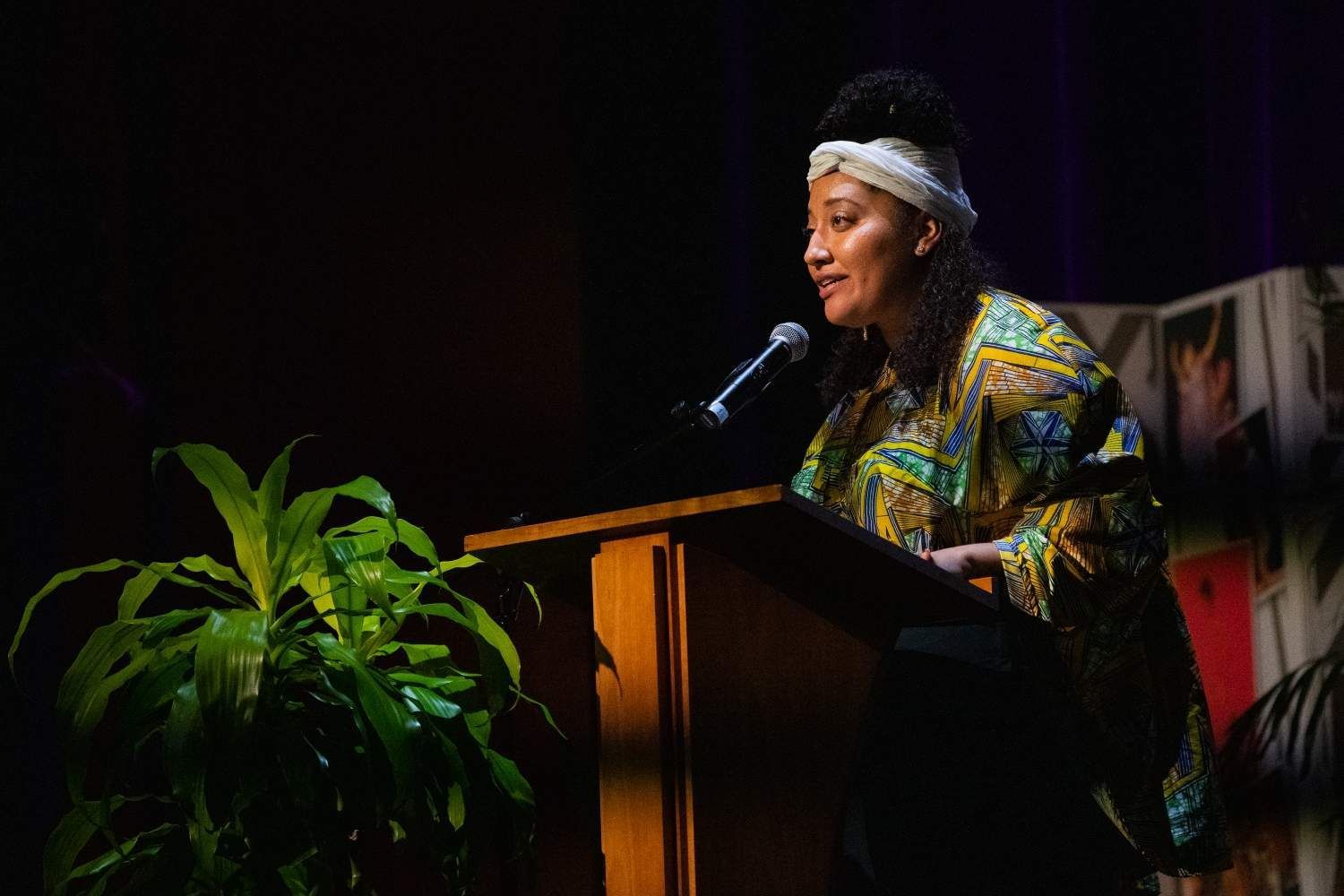 Woman in patterned shirt speaks at a podium.  She has a white head wrap and is near a leafy plant.
