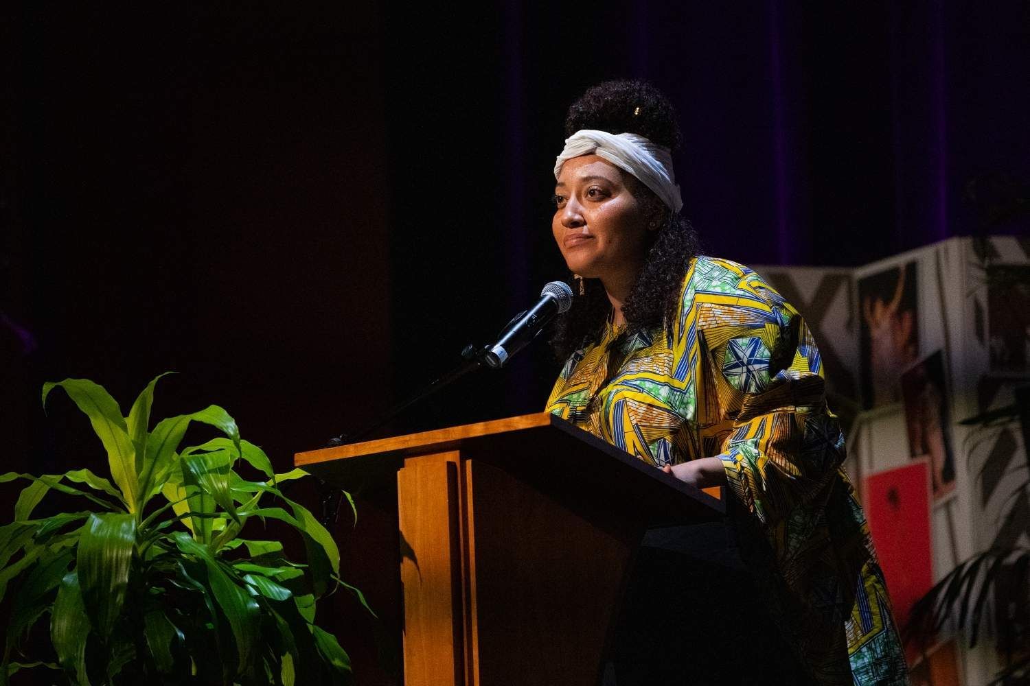 Woman at a podium speaking, wearing a patterned shirt and headscarf, indoors near a plant.