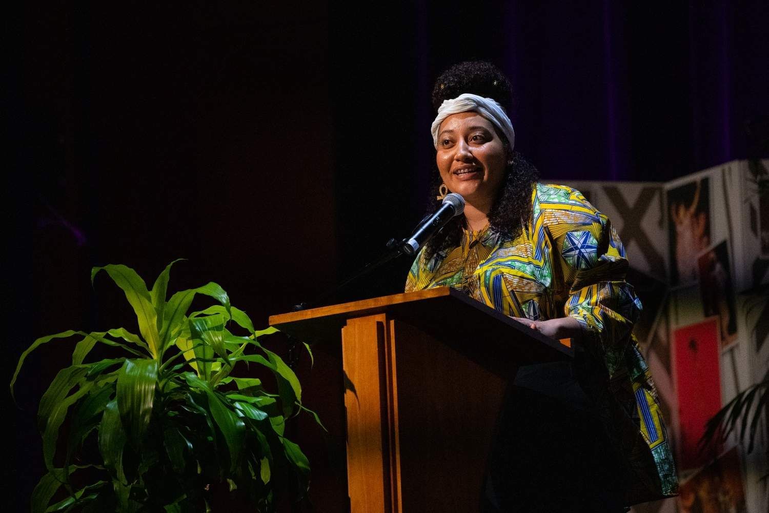 Woman speaking at a podium, wearing a patterned dress and headband. Indoor stage with plant.
