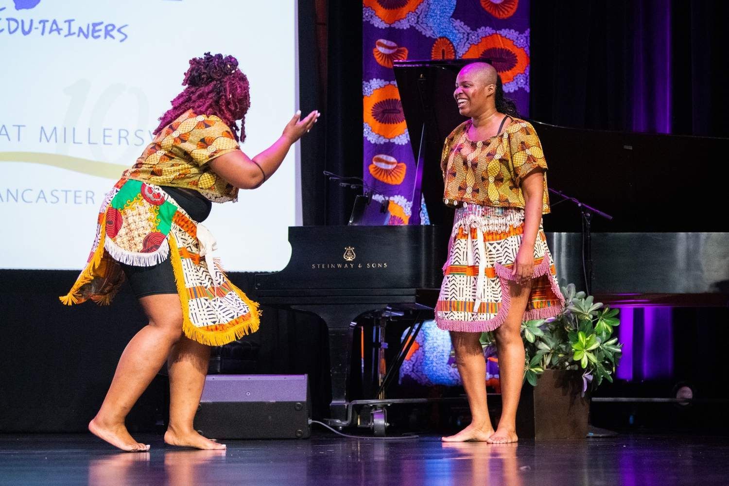 Two women on stage, wearing matching patterned outfits, interacting near a piano.