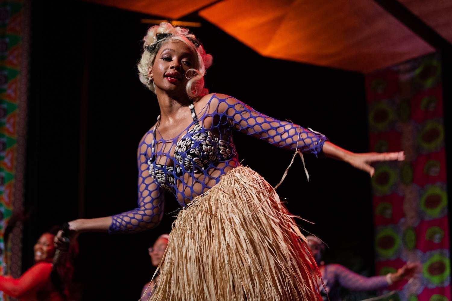 Dancer in blue netted top, shell bra, and straw skirt, performing onstage, arms outstretched.