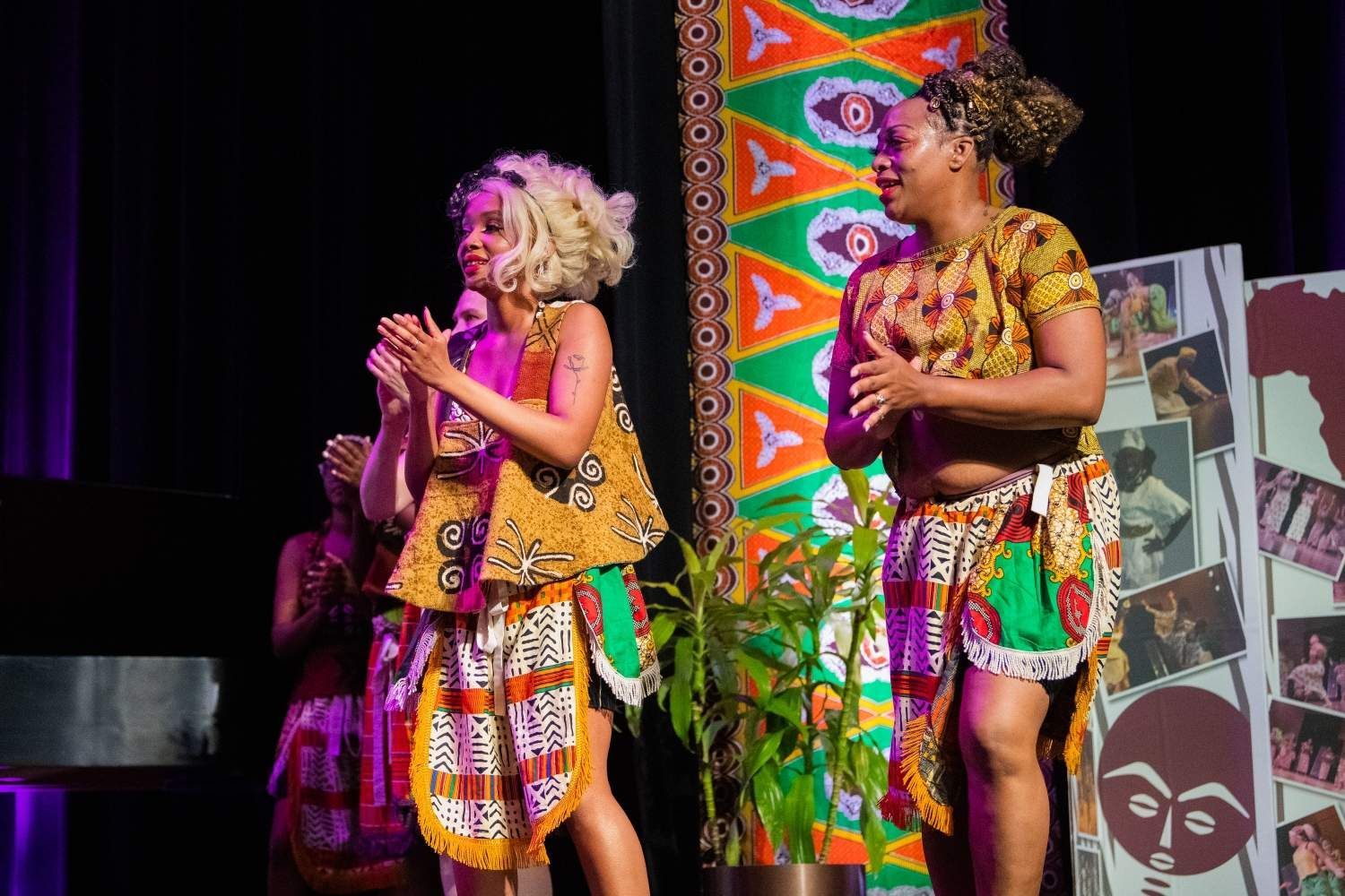 Two women in colorful African-inspired outfits clap on stage. A backdrop with tribal patterns.