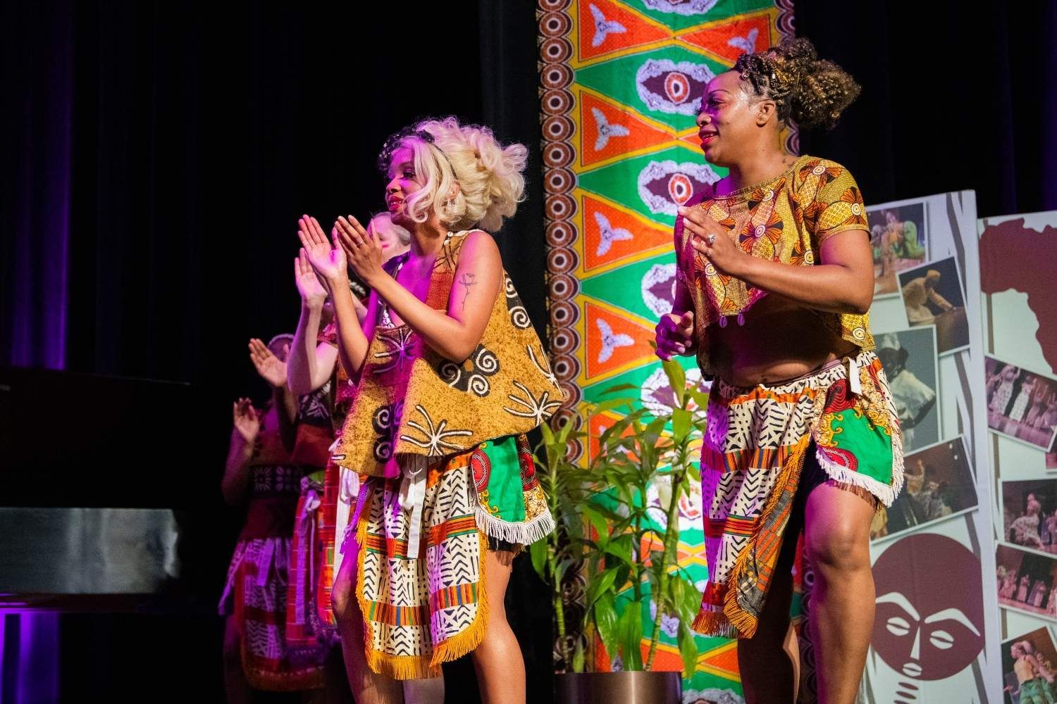 Women in African attire clap on stage, vibrant patterns, cultural performance.