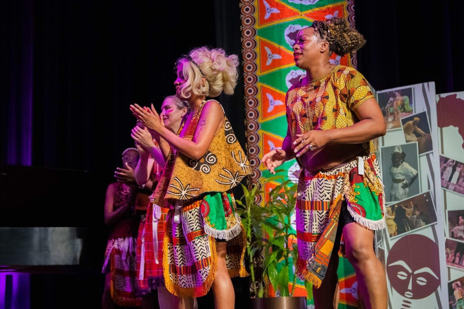 Stage performance: Two women in colorful African-style outfits onstage, with others applauding.