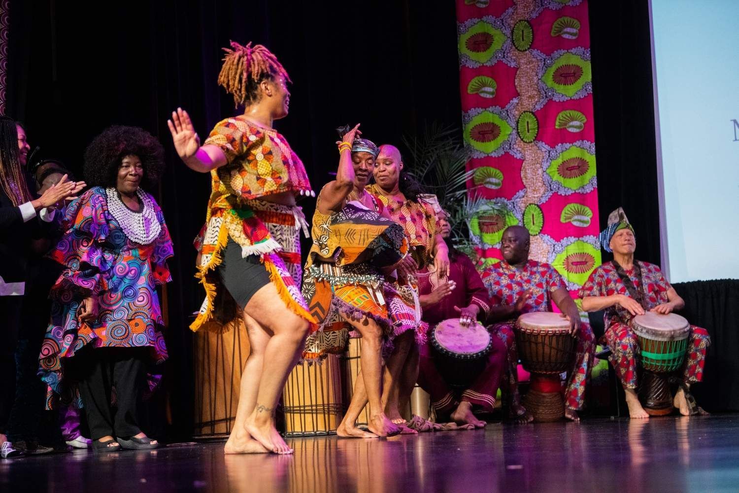People in colorful African attire perform on stage, including dancers and drummers.
