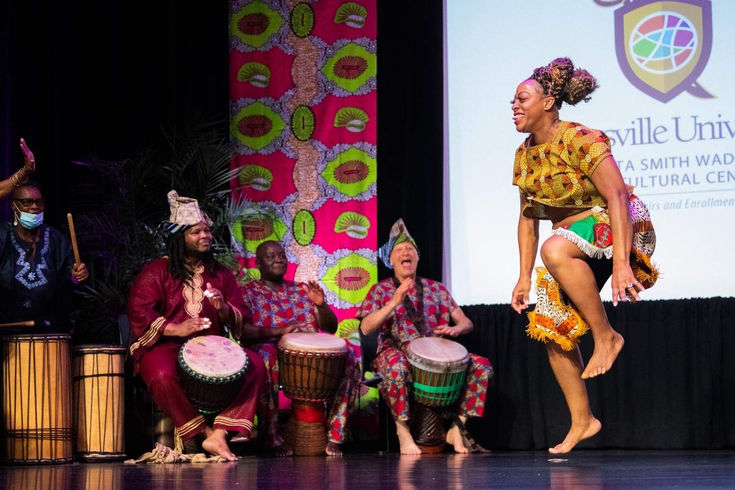 A woman dances barefoot on stage as a group plays drums. Vibrant African print decorates the backdrop.