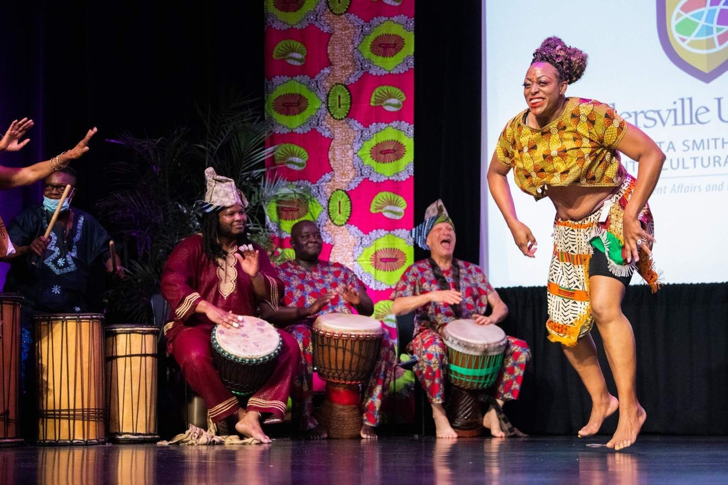 A woman dances joyfully on stage as a group of people play drums; stage is decorated in colorful patterns.