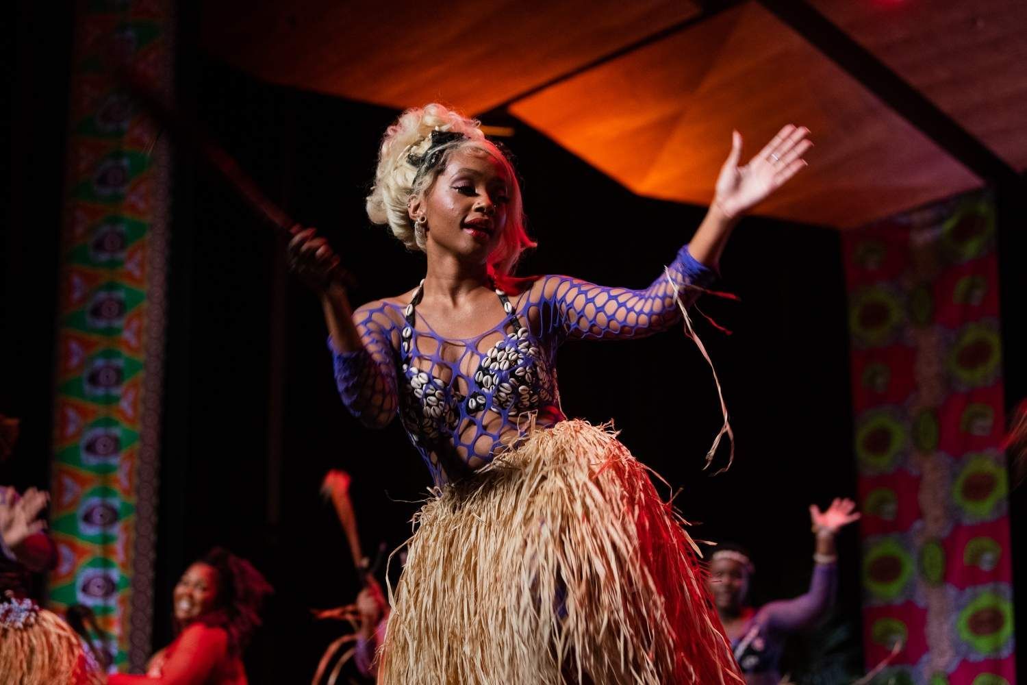 Woman dancing on stage, wearing a blue dress and straw skirt. Background with colorful patterns, warm lighting.