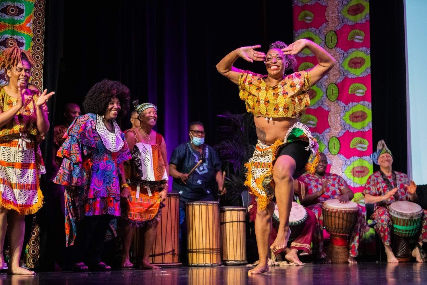African dancers in colorful outfits perform on stage with drummers.
