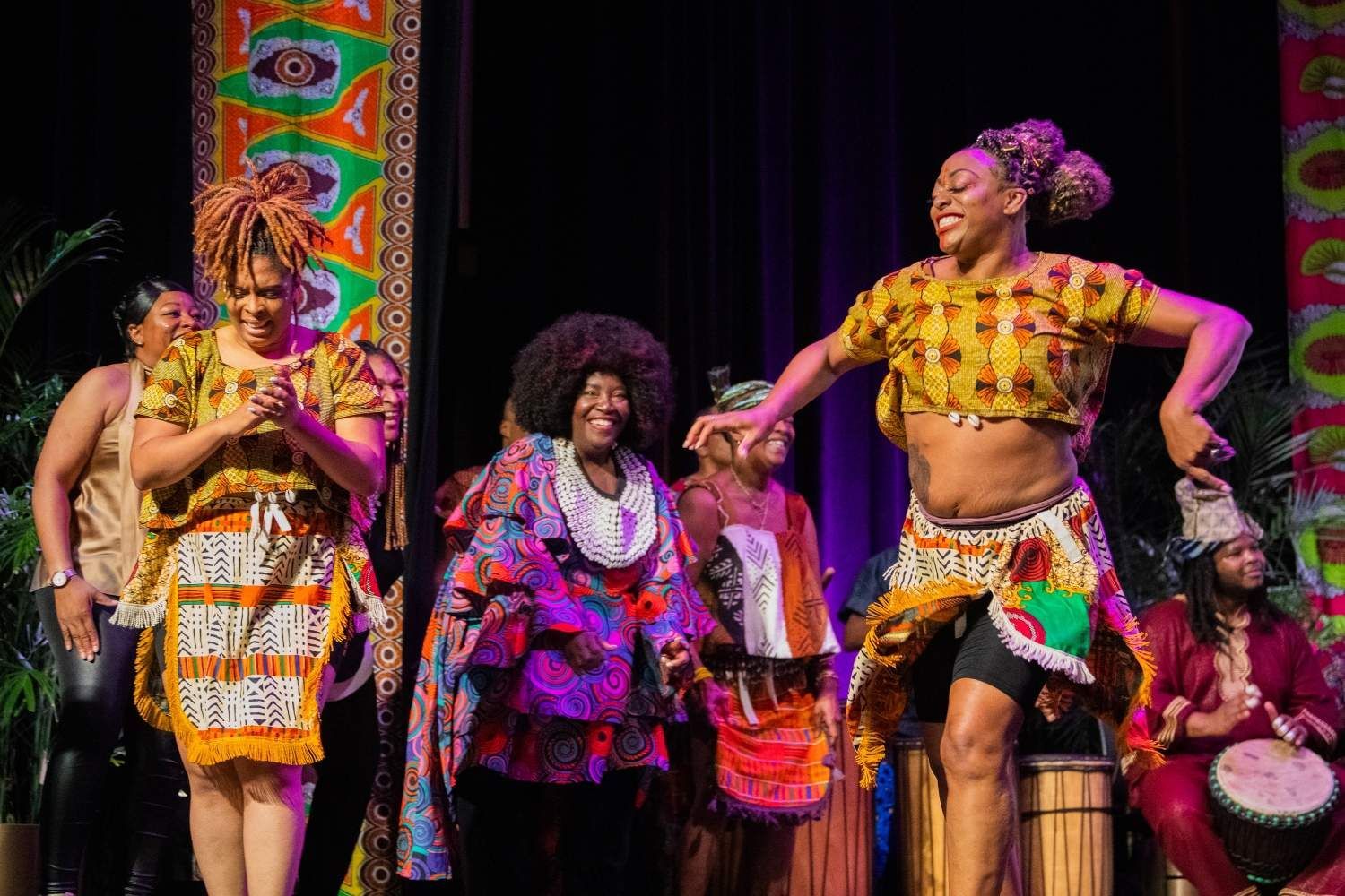African dancers on stage, vibrant fabrics, drum, joyful expressions, colorful backdrop.