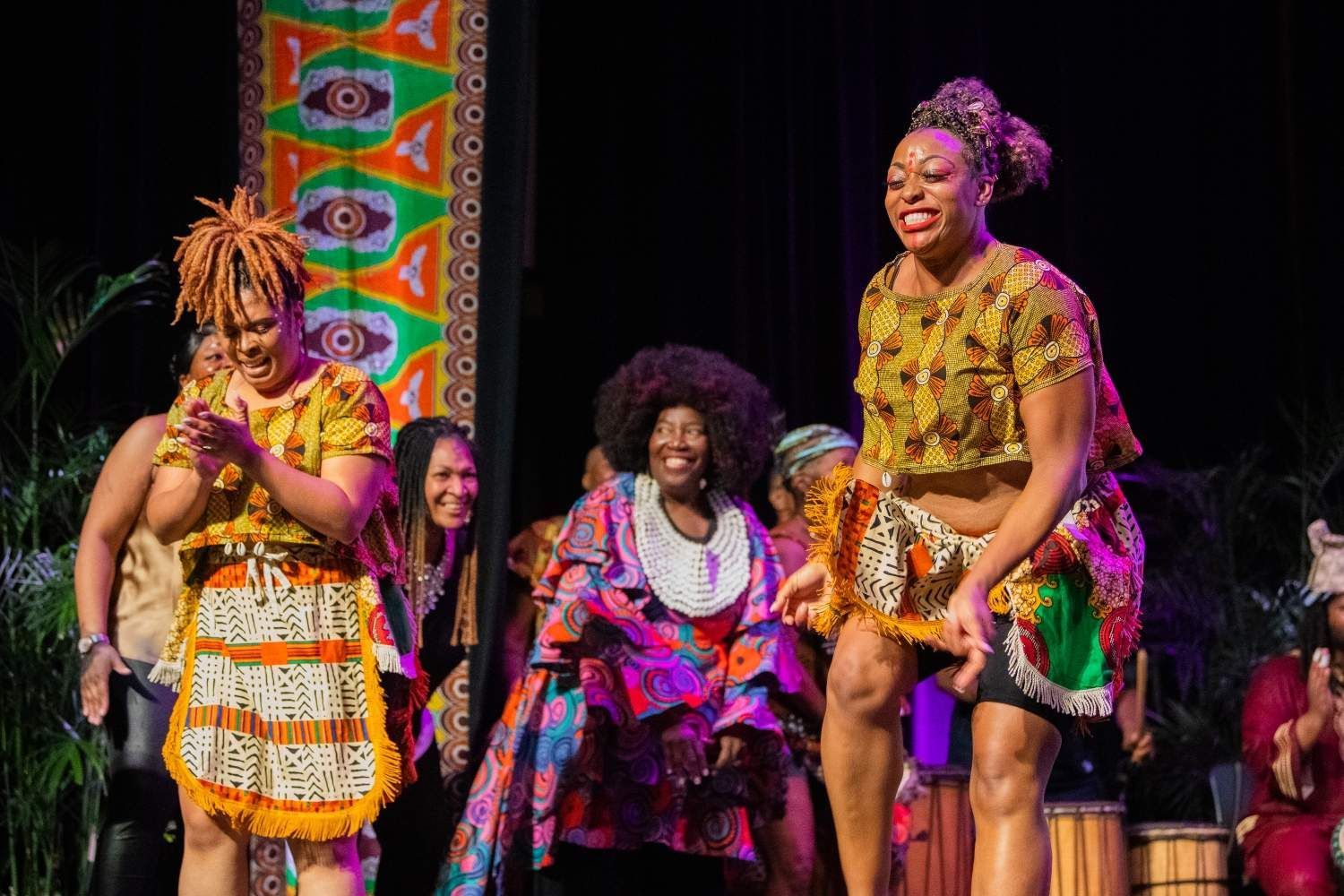 People on stage in vibrant African attire perform; smiling, clapping, and laughing.