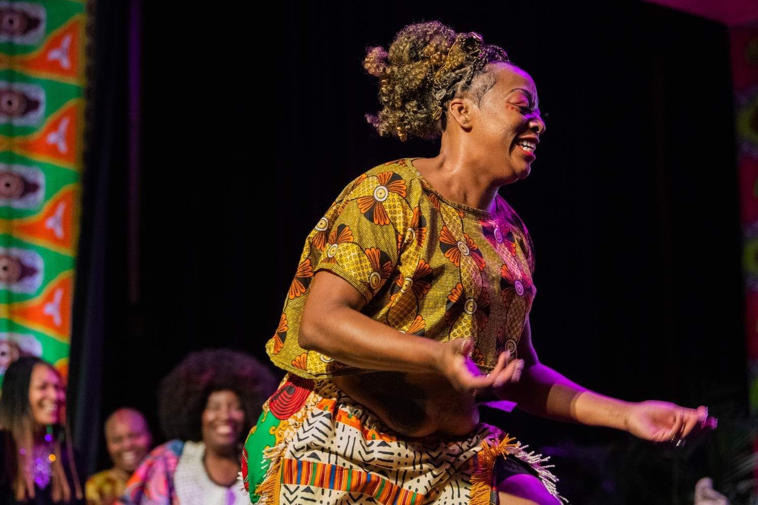 Woman dancing on stage wearing patterned African print clothing, smiling. Other women seated, watching.