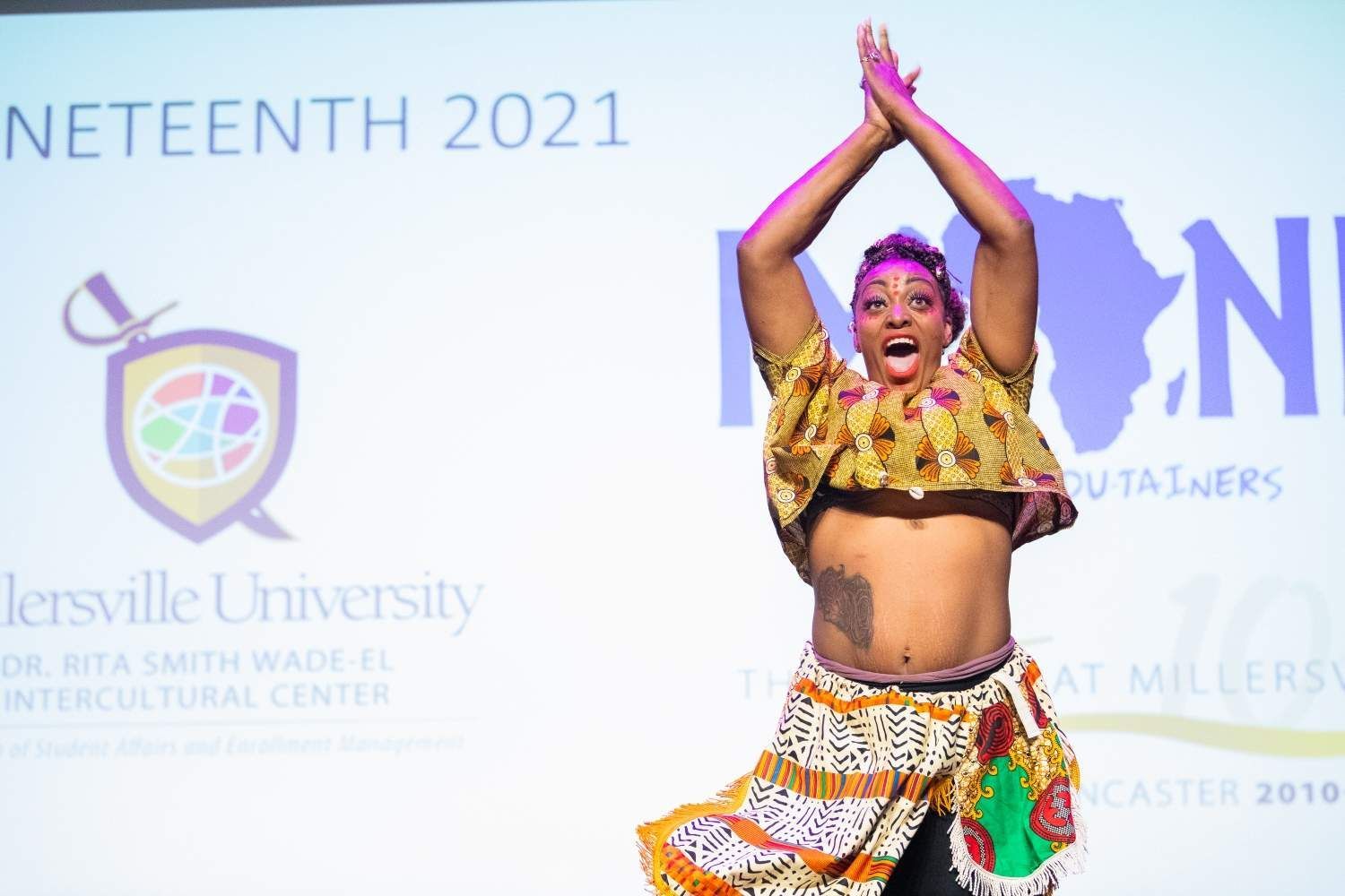 Woman in colorful African attire performs on stage for Juneteenth, arms raised. Millersville University backdrop.