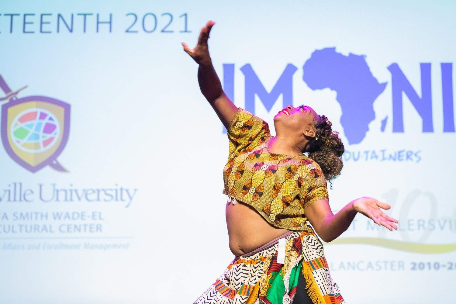 Woman dancing on stage with arms raised, African-themed attire, projected image of Africa in background.