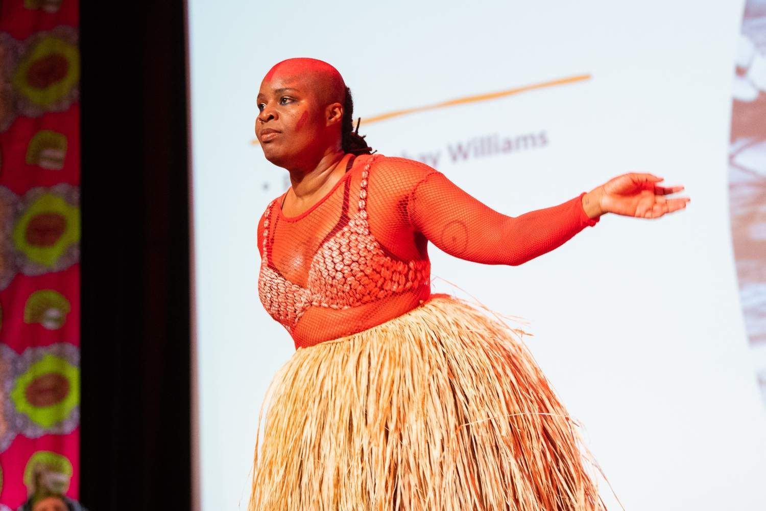 Woman in red mesh top and straw skirt on stage, arms outstretched, with a serious expression.