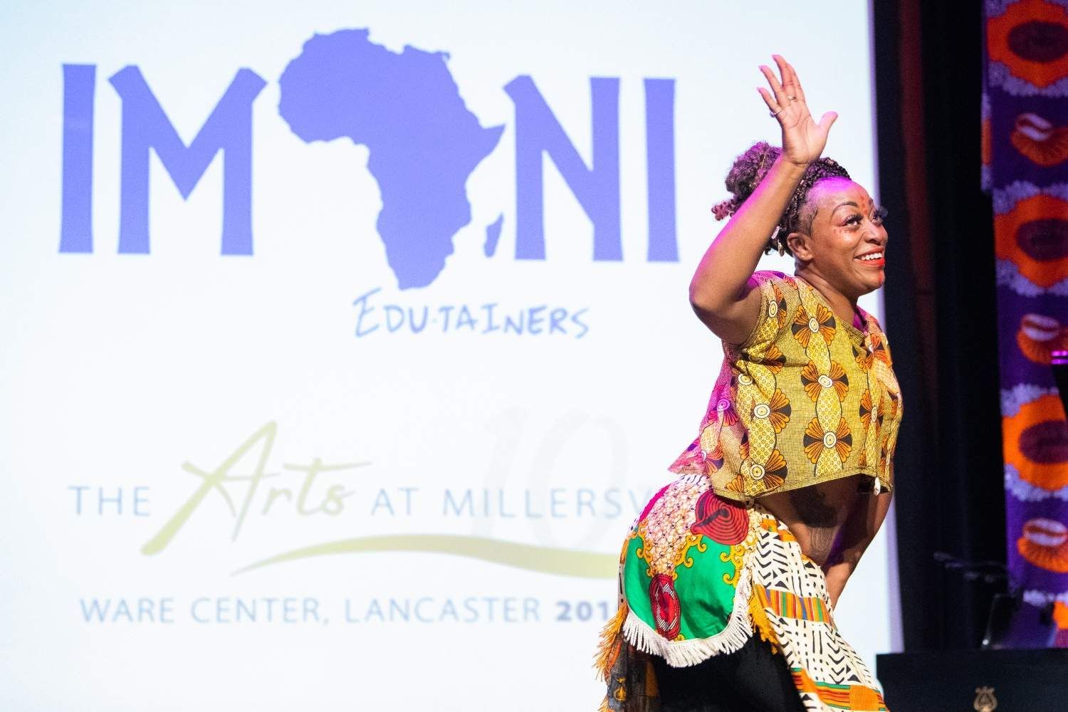 Woman in African print costume on stage, waving. 