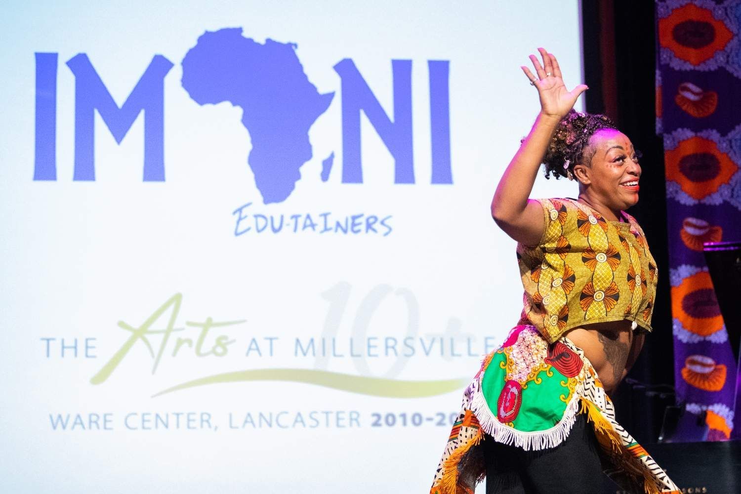 Woman in colorful African attire dancing onstage, smiling, in front of a screen with text and a map of Africa.