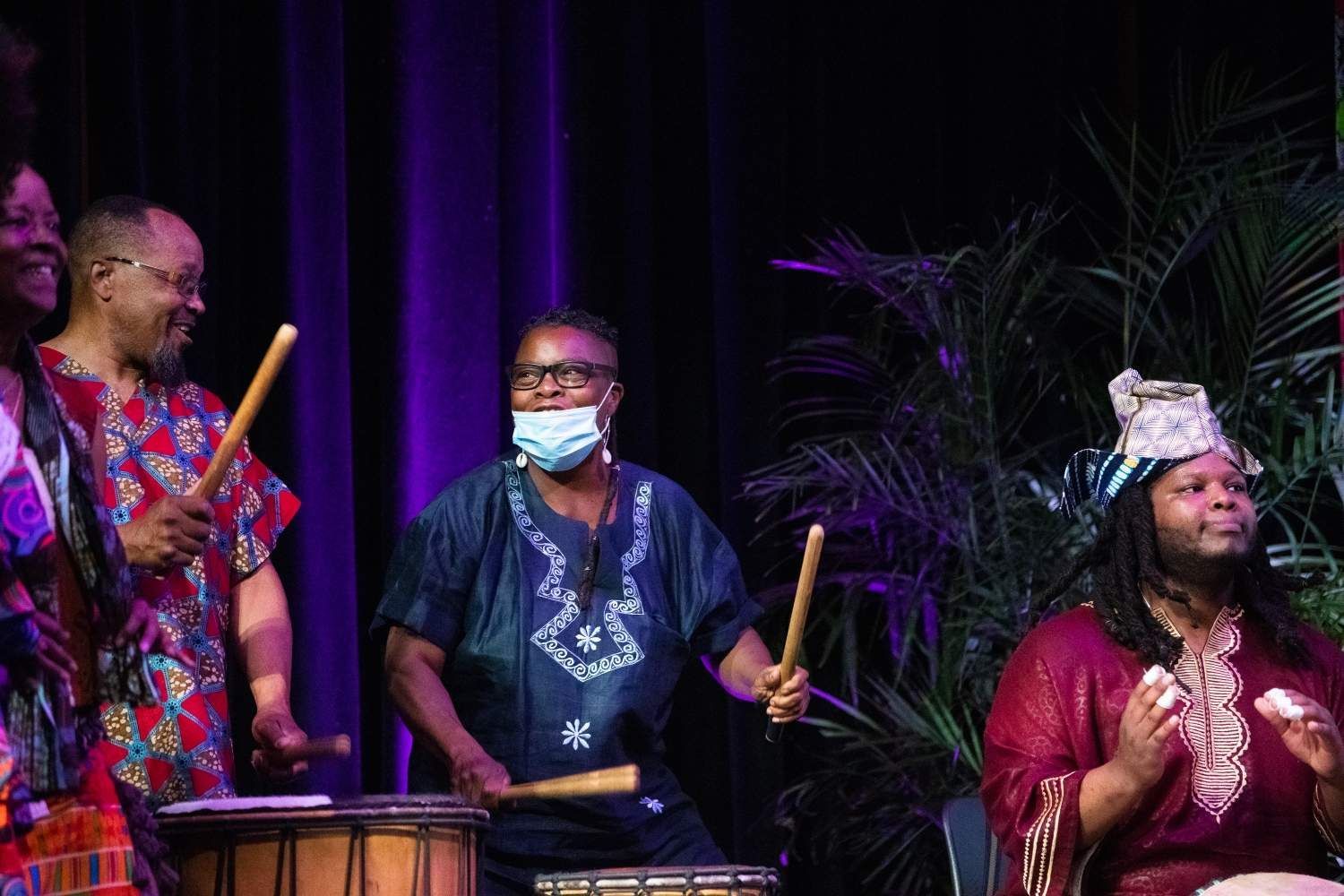 People in African attire play drums on a stage, one wearing a mask. Purple backdrop with greenery.