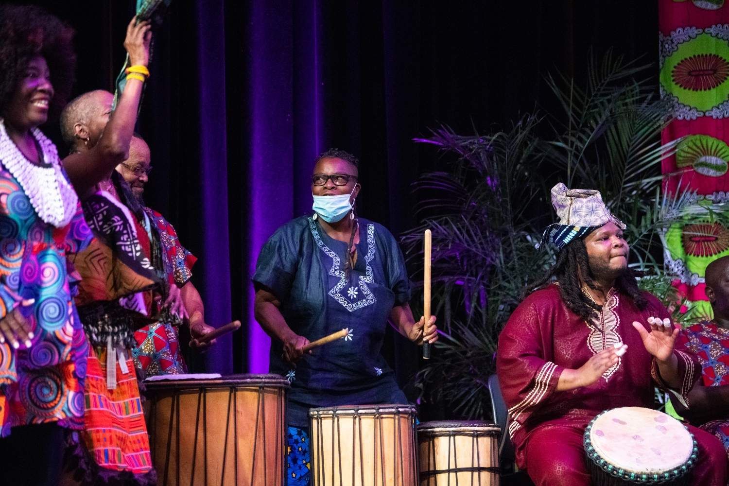 People in colorful African attire playing drums on stage; festive setting.