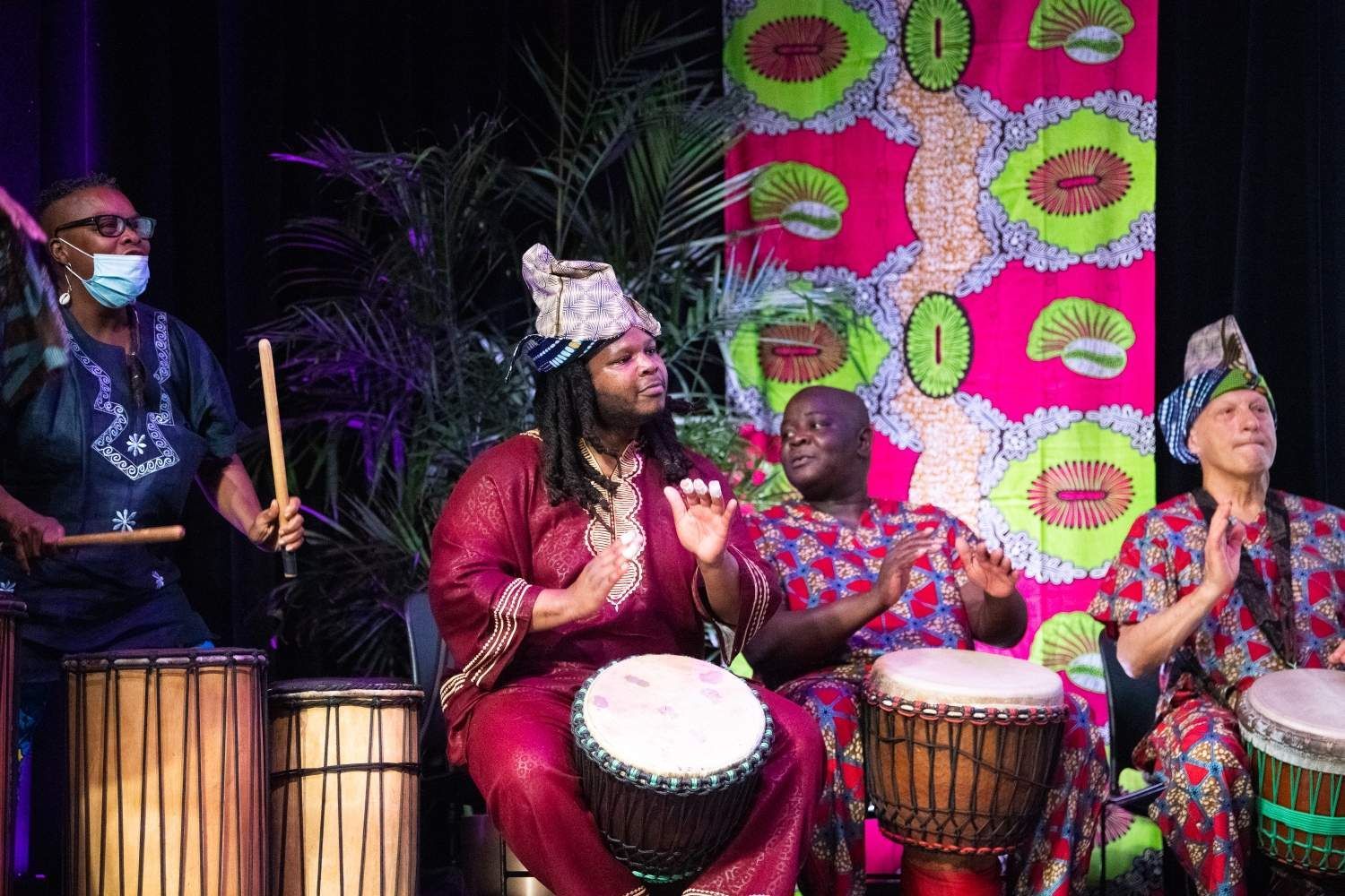 Group of four musicians playing drums, dressed in African-inspired clothing. Stage with colorful fabric backdrop.
